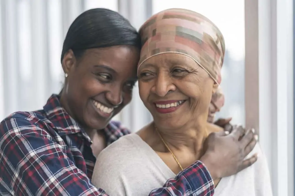 A black senior woman with cancer is wearing a scarf on her head. Her adult daughter is giving her a hug. Both women are smiling with gratitude and hope for recovery.