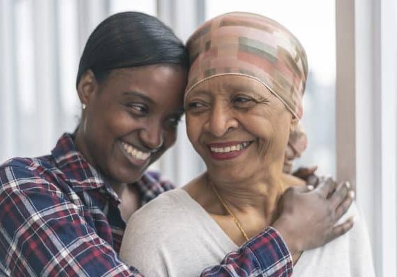 A black senior woman with cancer is wearing a scarf on her head. Her adult daughter is giving her a hug. Both women are smiling with gratitude and hope for recovery.