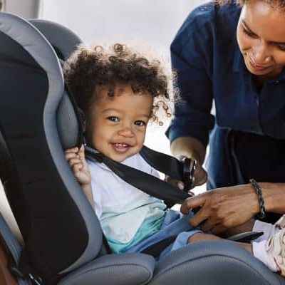 Side view of a happy little boy looking at camera while his mother buckling him in a car seat