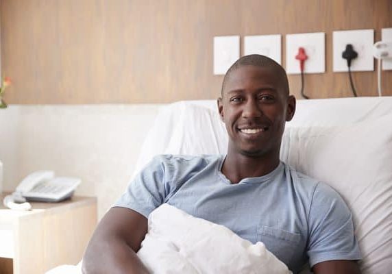 Portrait Of Male Patient Lying In Hospital Bed Smiling At Camera